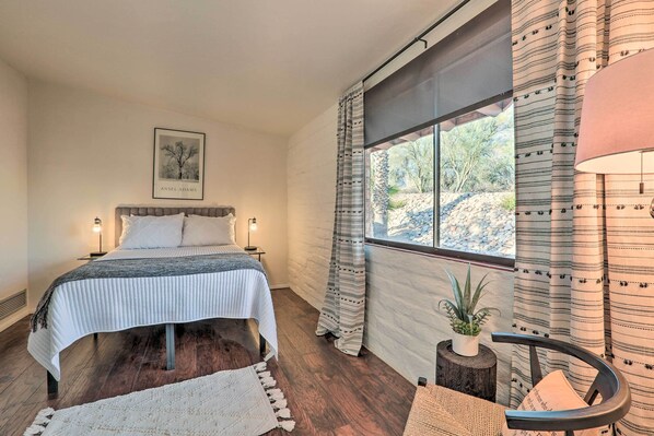 A bedroom with a bed, bedside lamps, a window with curtains, a framed picture on the wall, and a small potted plant.