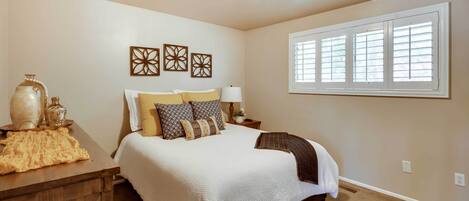 Un dormitorio con una cama grande, una mesita de noche, una lámpara, decoración en la pared y una ventana con persianas.