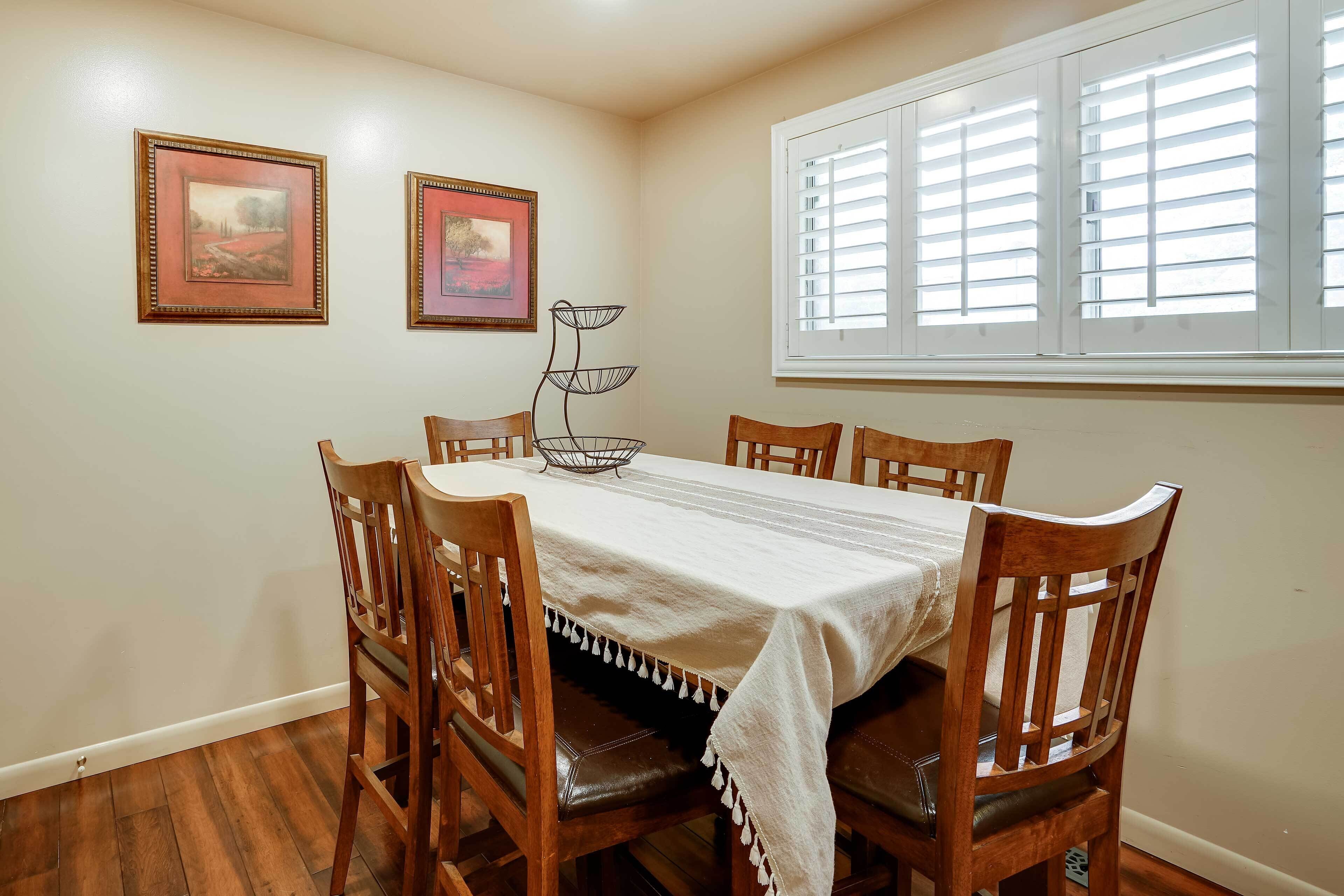 A dining area with a table set for six, surrounded by wooden chairs. There are two framed artworks on the wall, a window with blinds, and a floor lamp.