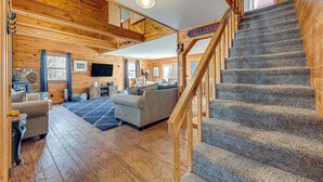 Un intérieur de chalet chaleureux avec un escalier, des murs en bois, un espace de vie comprenant un canapé et un téléviseur, et une enseigne indiquant « SKIL LODGE ».