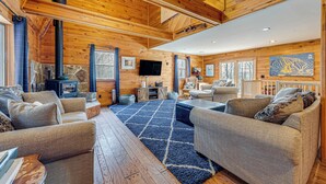Un intérieur de chalet chaleureux avec des murs en bois, une cheminée en pierre, un grand téléviseur et des sièges confortables.