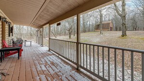 Une véranda couverte avec un plancher en bois, une balustrade et une vue sur une zone boisée, avec un petit bâtiment visible au loin.