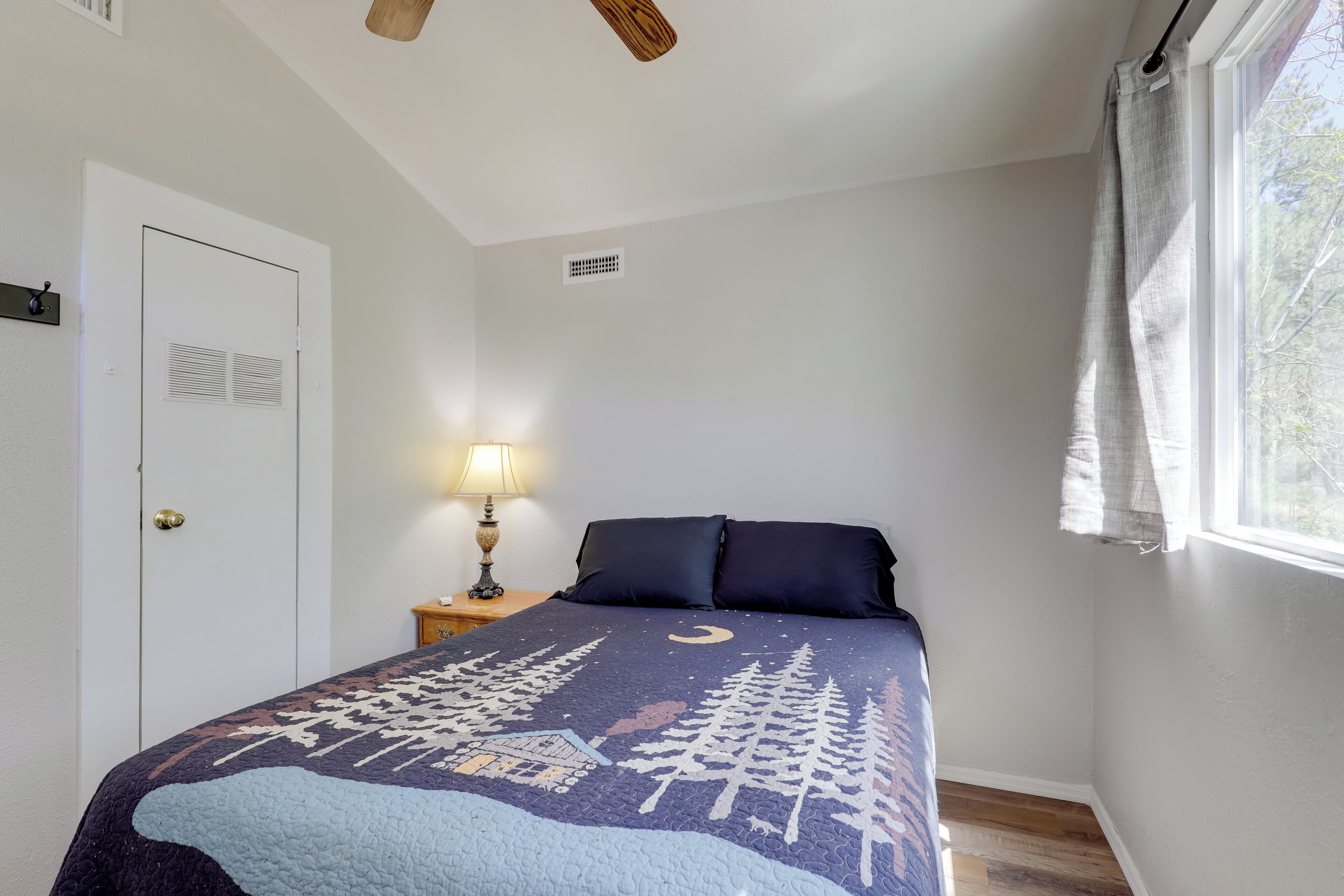 A bedroom with a bed, a nightstand, a lamp, a window with curtains, and a door with a lock.