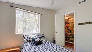 Een slaapkamer met een geruit bedovertrek, een houten dressoir, een raam met uitzicht op bomen, en een deur die leidt naar een kamer met een stenen muur en kunstwerken.