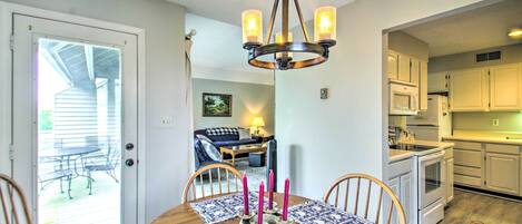 Uma sala de jantar com mesa de madeira, cadeiras e um lustre. Cozinha visível ao fundo, com armários e eletrodomésticos brancos.