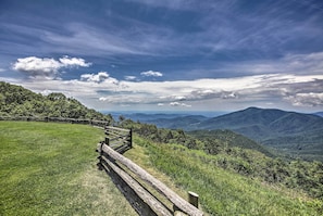 Une clôture en bois longe un sentier bordé d’herbe, offrant une vue sur des montagnes lointaines et un ciel partiellement nuageux.