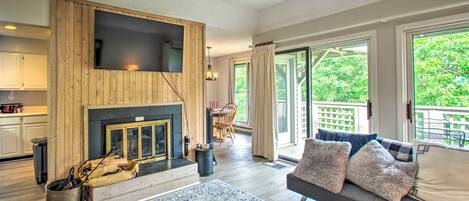 Una sala moderna con chimenea, un televisor de pantalla plana montado en una pared de madera, un sofá con almohadones decorativos y grandes ventanas que ofrecen vistas al exterior.