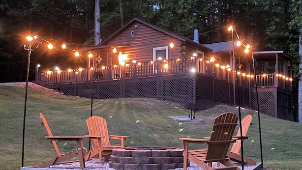 En stuga av trä med en veranda, omgiven av ett staket, och med två Adirondack-stolar och en eldstad.