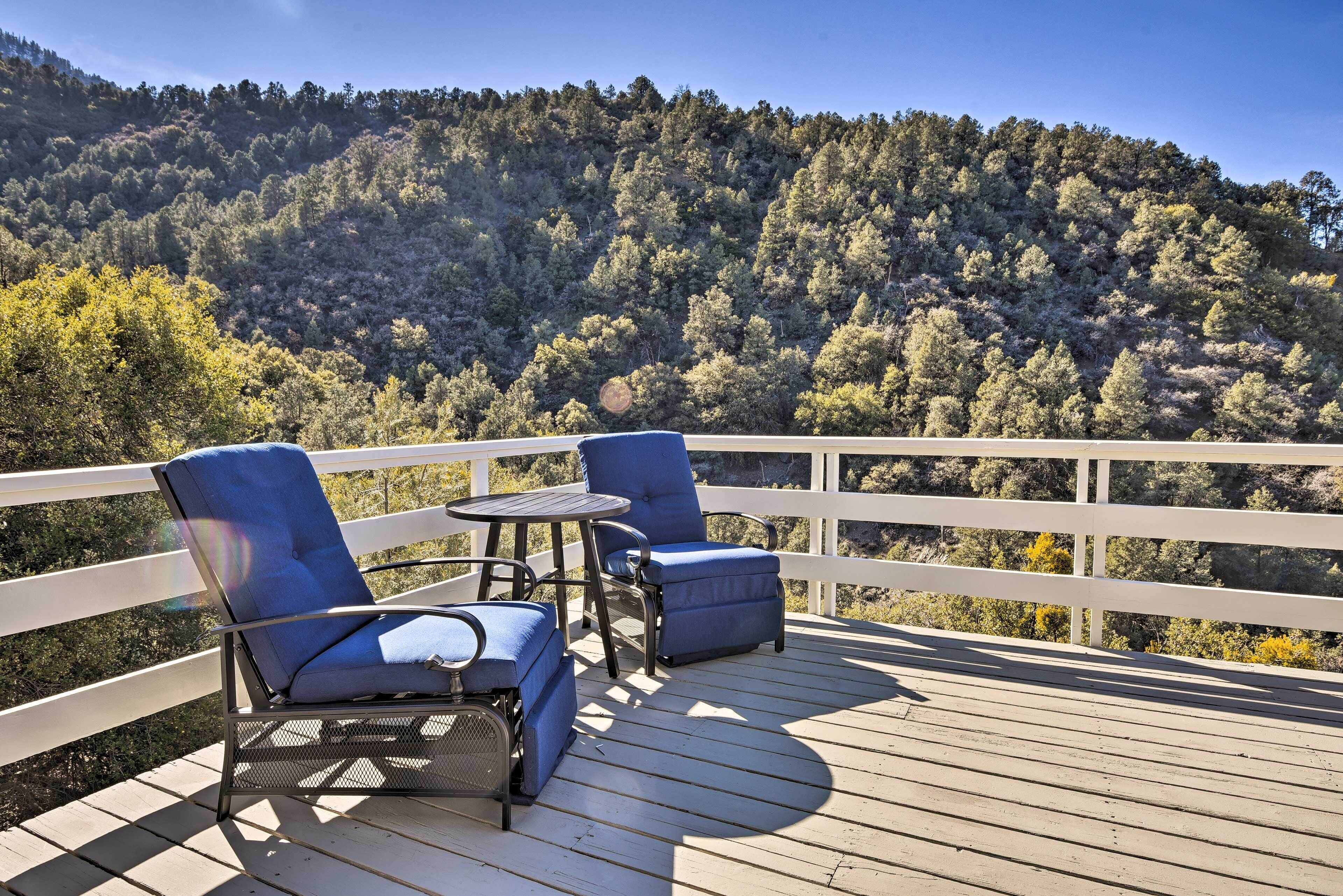 A deck with two blue chairs and a small table, overlooking a forested hillside.