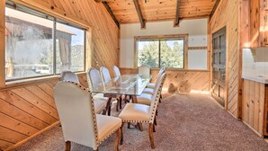 A wooden interior with a glass-top dining table surrounded by six chairs, large windows, and a chandelier. - Luxe Escape w/ Decks, Mtn Views, Game Room! (Pine Mountain Club)