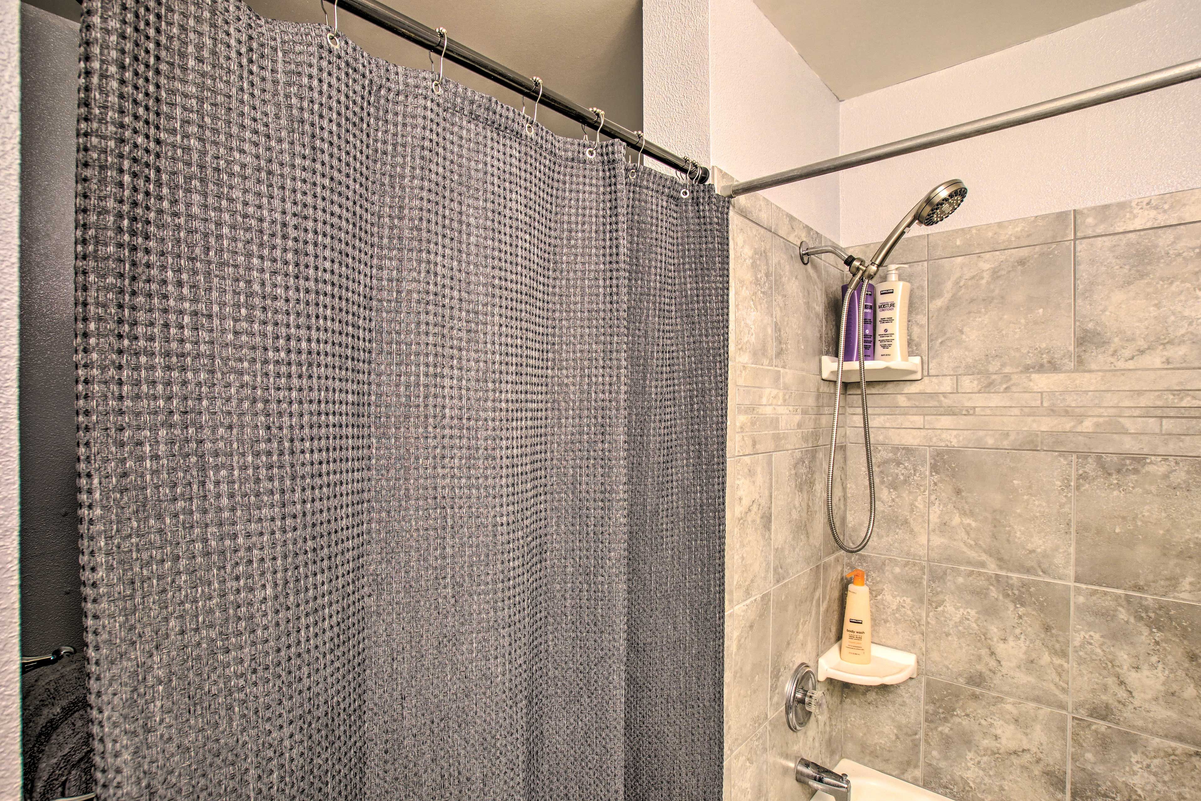 Un espace douche avec un rideau de douche gris, une douchette à main et un distributeur de savon placé sur une étagère.