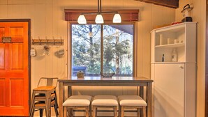 A kitchen with a wooden table, three bar stools, a white refrigerator, and a window with a view of trees.
