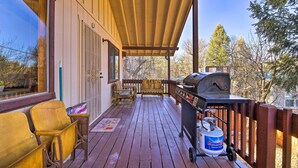 A wooden deck with a grill, chairs, and a window overlooking a wooded area.
