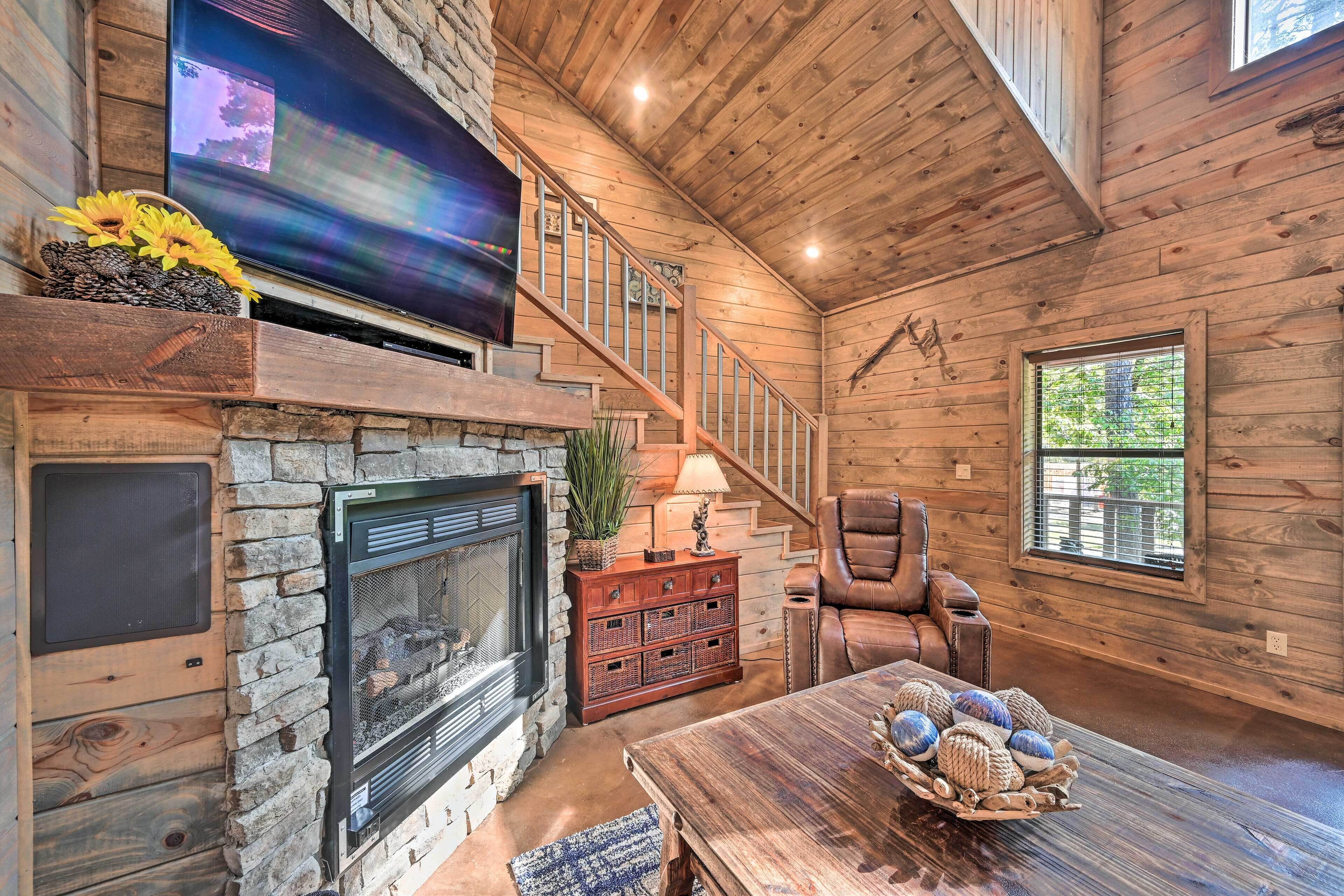 A cozy cabin interior with a stone fireplace, a wooden staircase, a leather armchair, and a large TV mounted on the wall.