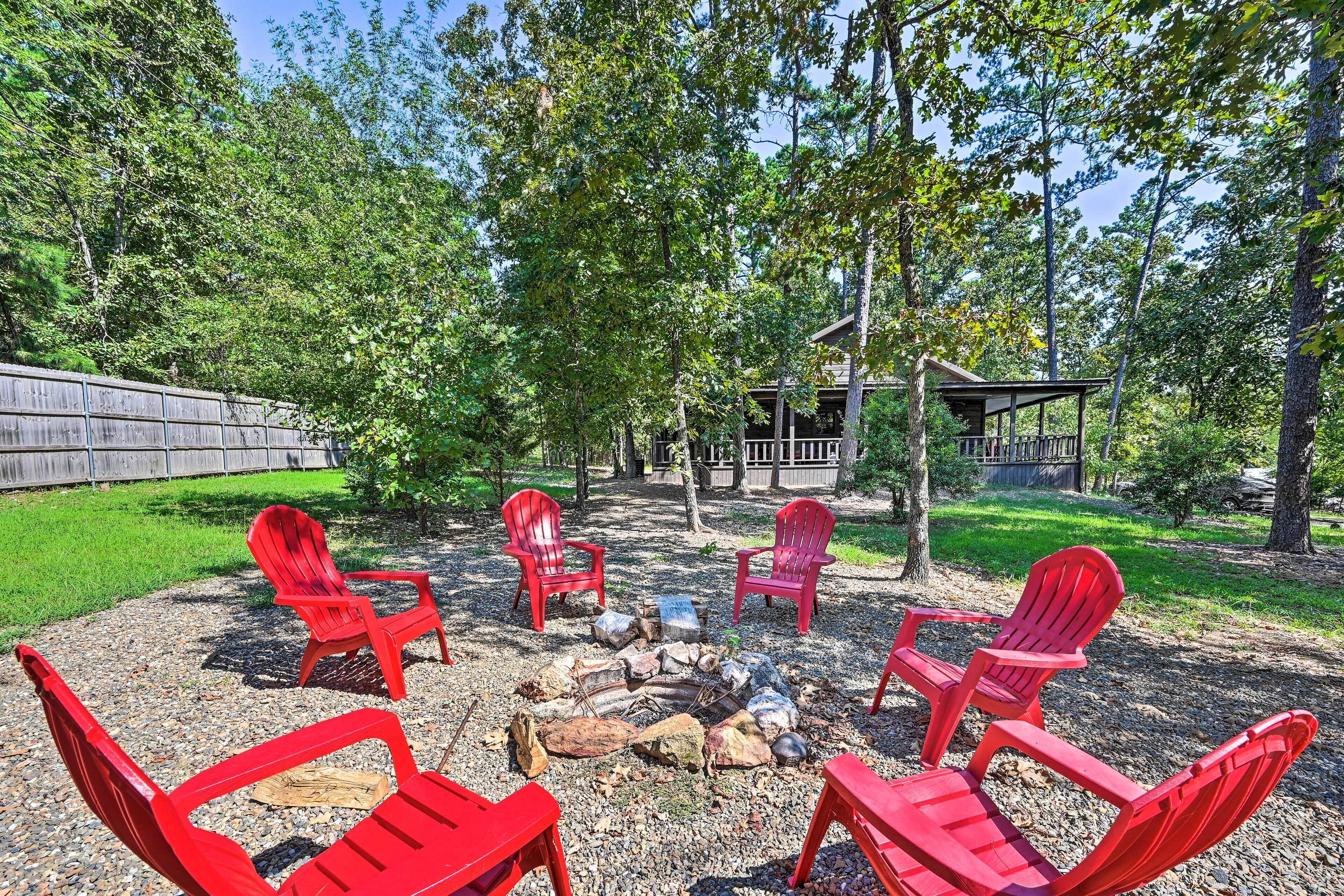 Ein Lagerfeuer, umgeben von roten Adirondack-Stühlen in einer bewaldeten Außenanlage mit einer Holzterrasse und einem Zaun im Hintergrund.