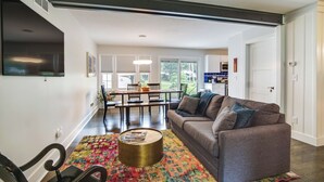 Una sala moderna con un sofá gris, una mesa de centro redonda y un tapete colorido. Se observa un comedor con una mesa y sillas, y una cocina al fondo.