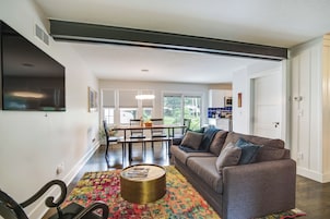 A modern living room with a gray sofa, a round coffee table, and a colorful rug. There is a dining area with a table and chairs, and a kitchen in the background.