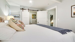 Un dormitorio con una cama grande, una mesita de noche con lámpara, una puerta que da al baño y una escalera.
