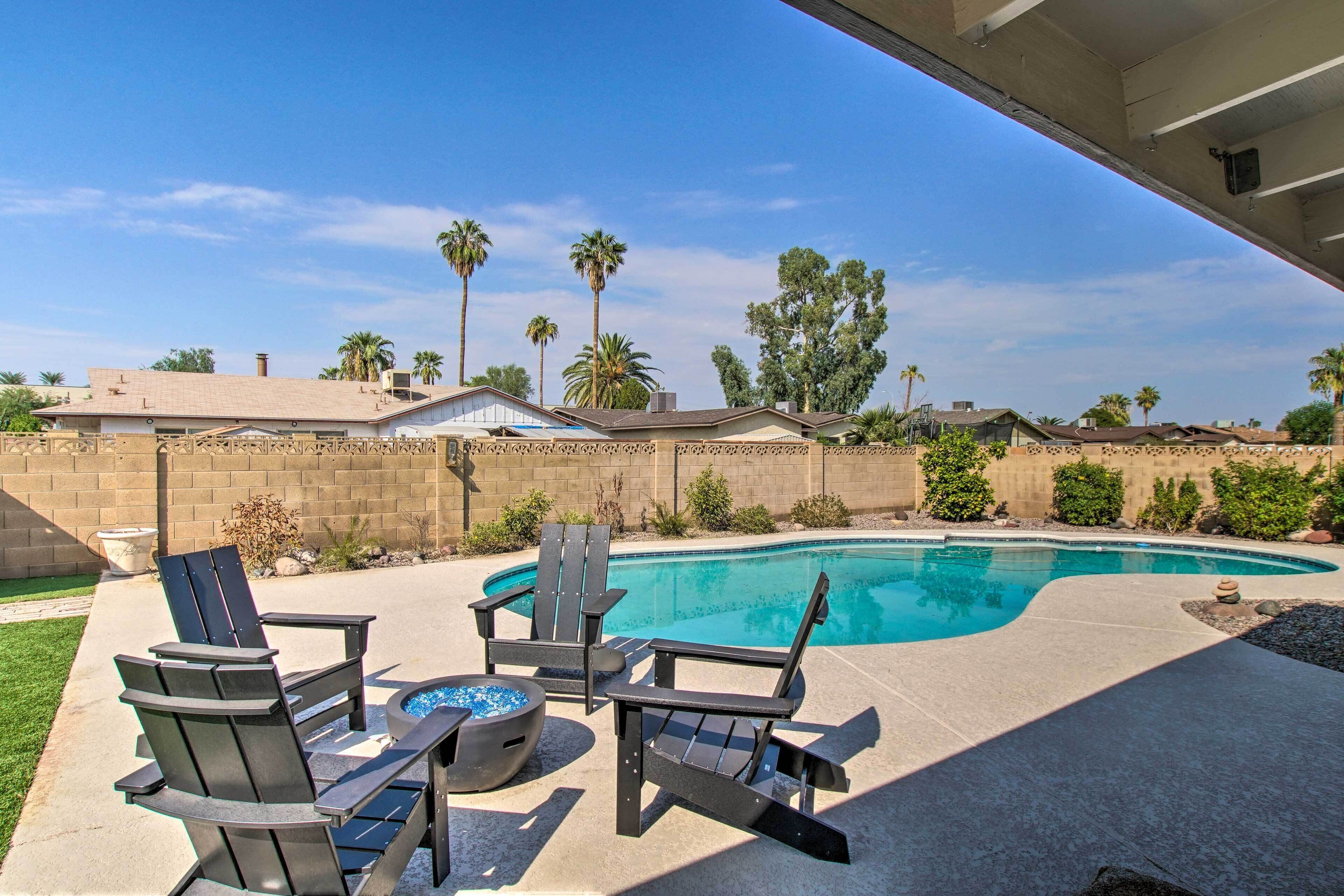 A backyard with a swimming pool, three Adirondack chairs, and a fire pit.