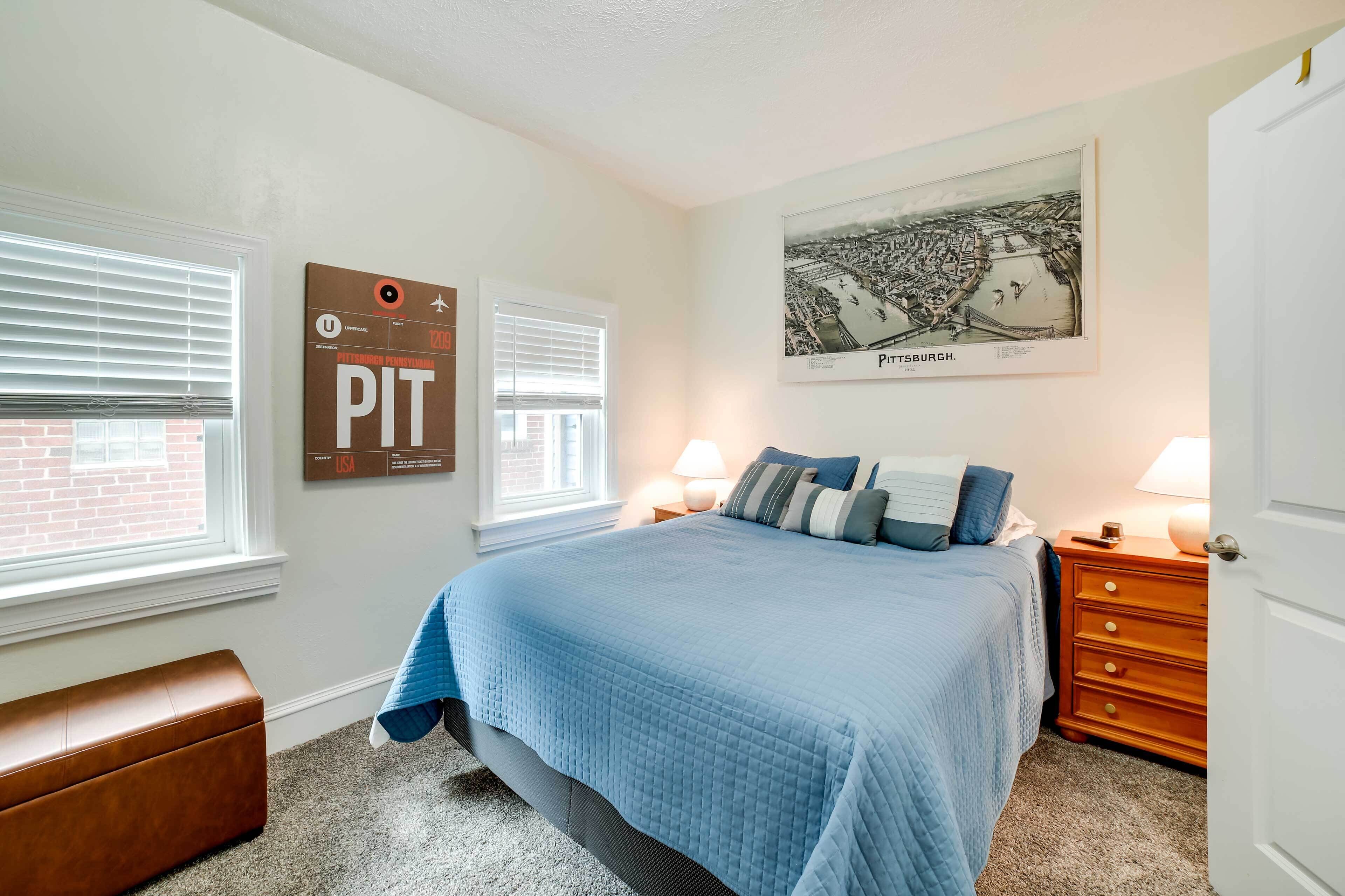 Une chambre à coucher comprenant un lit, deux tables de chevet, une commode, un banc et un mur orné d’un tableau encadré et d’une affiche.