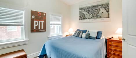 Une chambre à coucher comprenant un lit, deux tables de chevet, une commode, un banc et un mur orné d’un tableau encadré et d’une affiche.