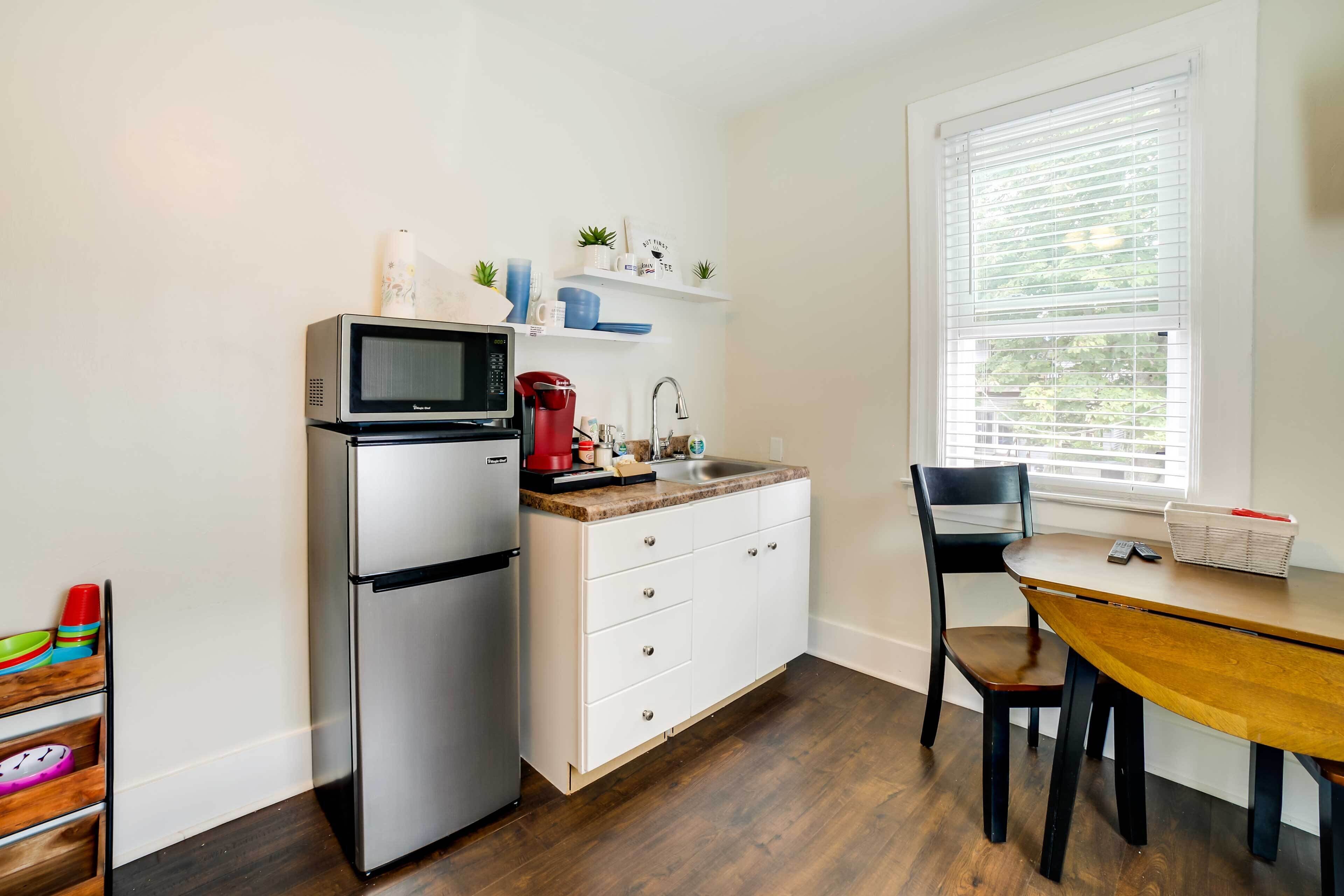 A compact kitchen with a refrigerator, microwave, and coffee maker. There is a wooden dining table with chairs, a window with blinds, and a small shelf with decorative items.