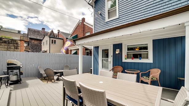 A covered patio with a wooden table and wicker chairs, a barbecue grill, and a view of neighboring houses. - Walk to Walnut St: Pet-friendly Pittsburgh Home! (Pittsburgh)