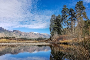 A calm river with a clear reflection, surrounded by trees and mountains under a partly cloudy sky.