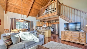 A spacious living area with a wooden ceiling, a sofa with cushions, a TV on a wooden cabinet, and a kitchen in the background. - Pet-friendly Munds Park Retreat w/ Wraparound Deck (Munds Park)
