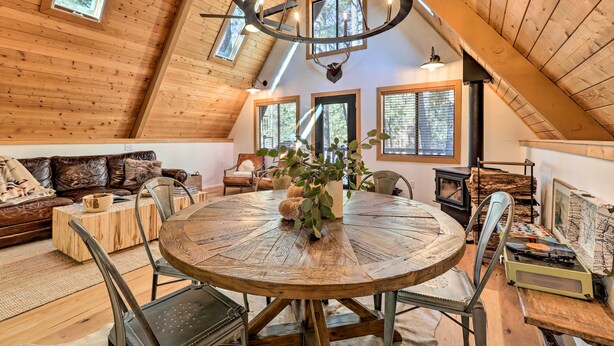A cozy loft-style room with a wooden ceiling, a round wooden dining table, a fireplace, and a large window with a view of trees. - Deck & Forest Views: Munds Park A-frame Cabin (Munds Park)