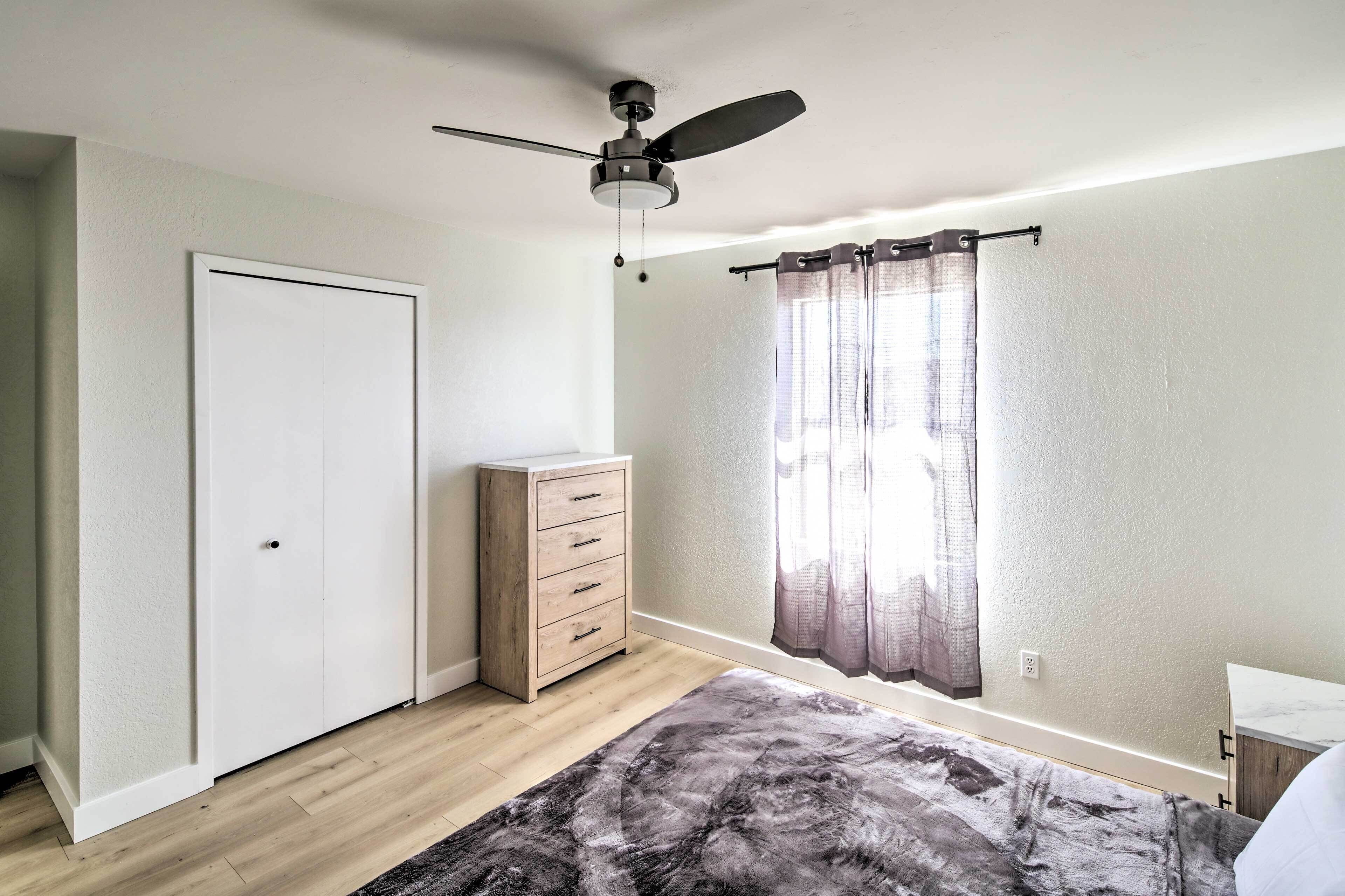 A bedroom with a ceiling fan, a wardrobe, a bedside table, a window with curtains, and a patterned rug.