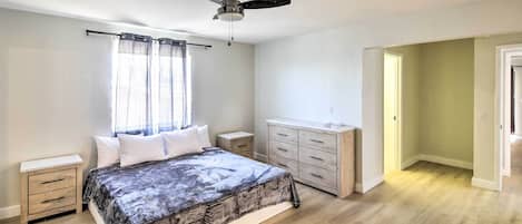 Een slaapkamer met een bed, nachtkastje, dressoir, plafondventilator en een raam met gordijnen.