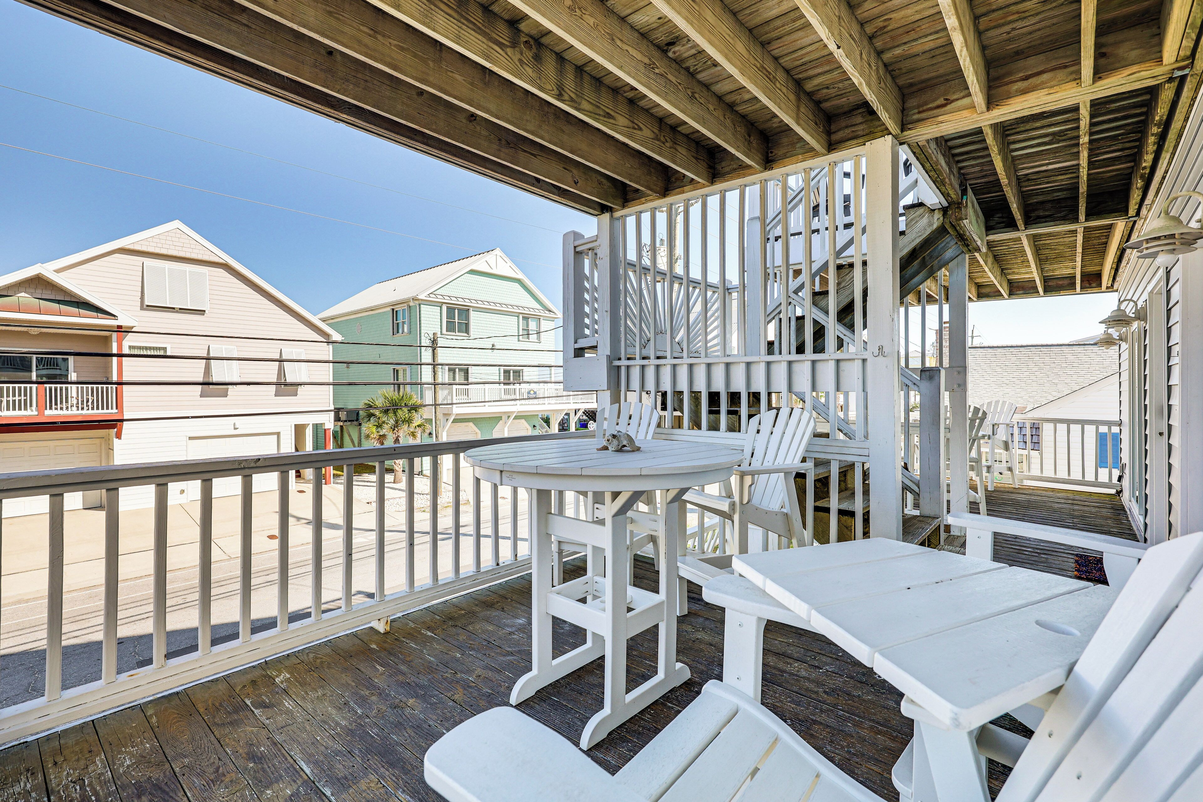 A wooden deck with white furniture, including a table and chairs, overlooking a street with houses.