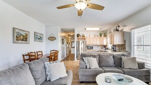 Ein modernes Wohnzimmer mit grauen Sofas, einem weißen Couchtisch und einem Deckenventilator. Im Hintergrund ist eine Küche mit hellen Schränken und einem Edelstahlkühlschrank zu sehen.