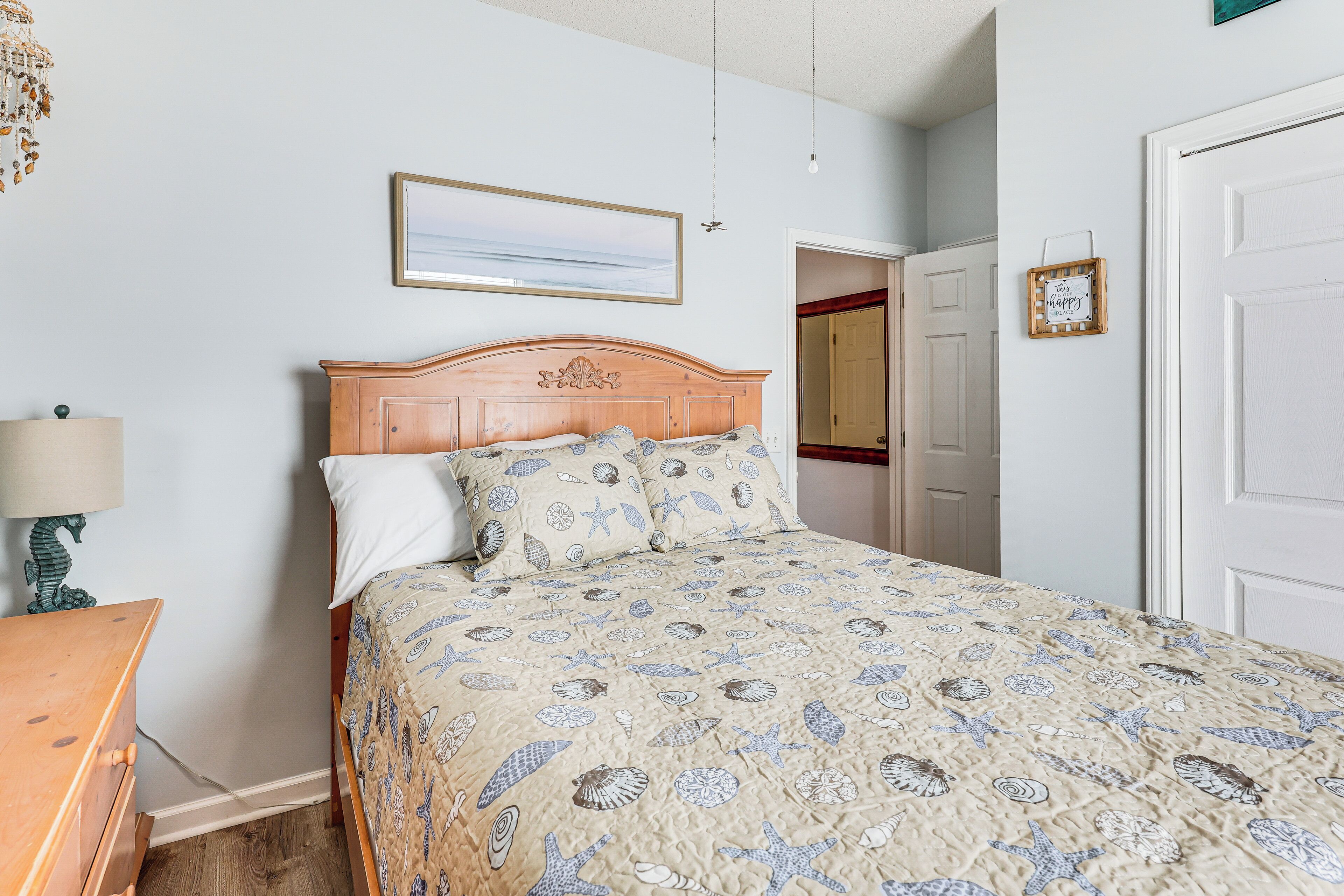 A bedroom with a wooden headboard, a bed with a patterned bedspread, a bedside table with a lamp, and a framed picture on the wall.