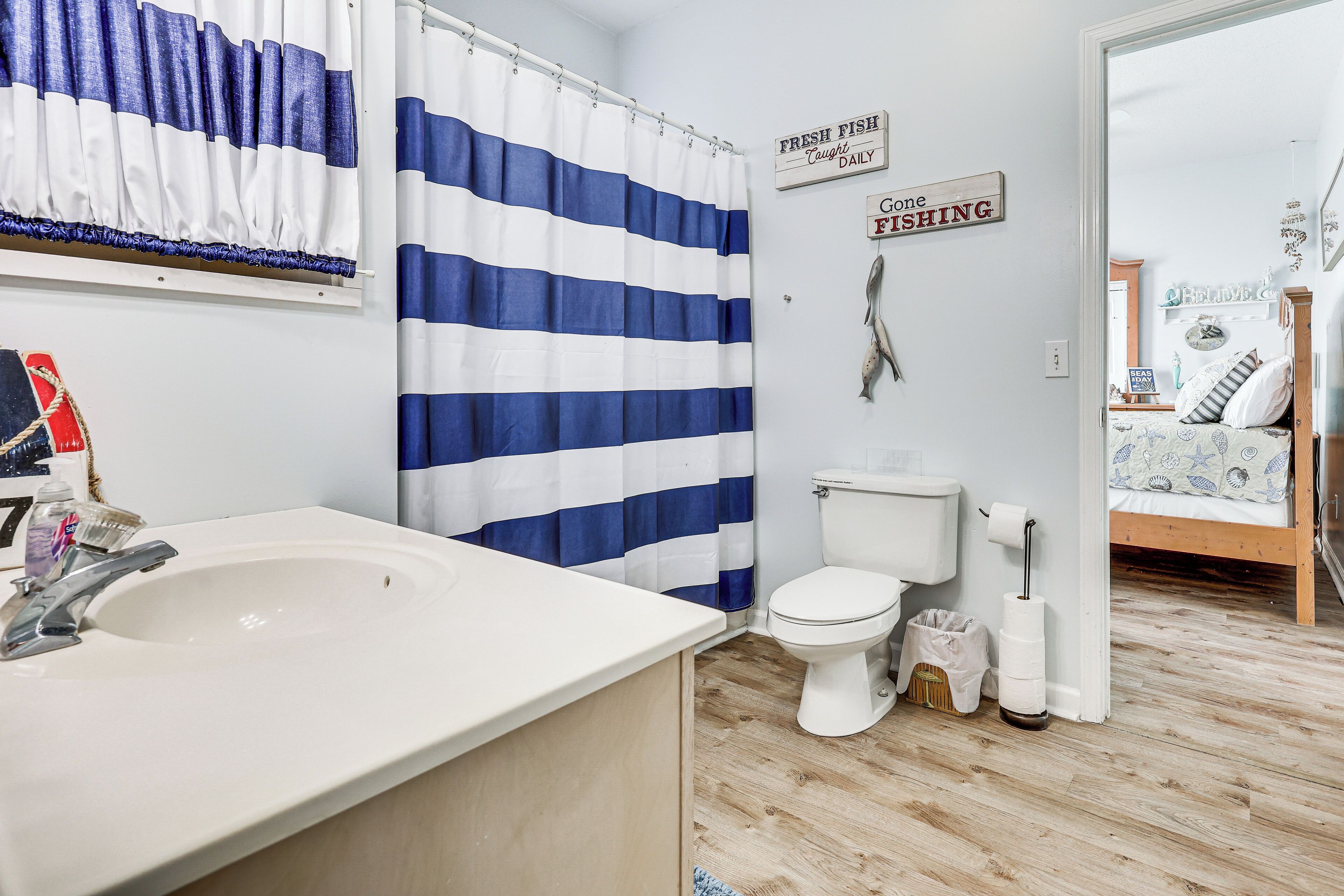 A bathroom with a striped shower curtain, a toilet, a sink, and a bed visible in the adjacent room.