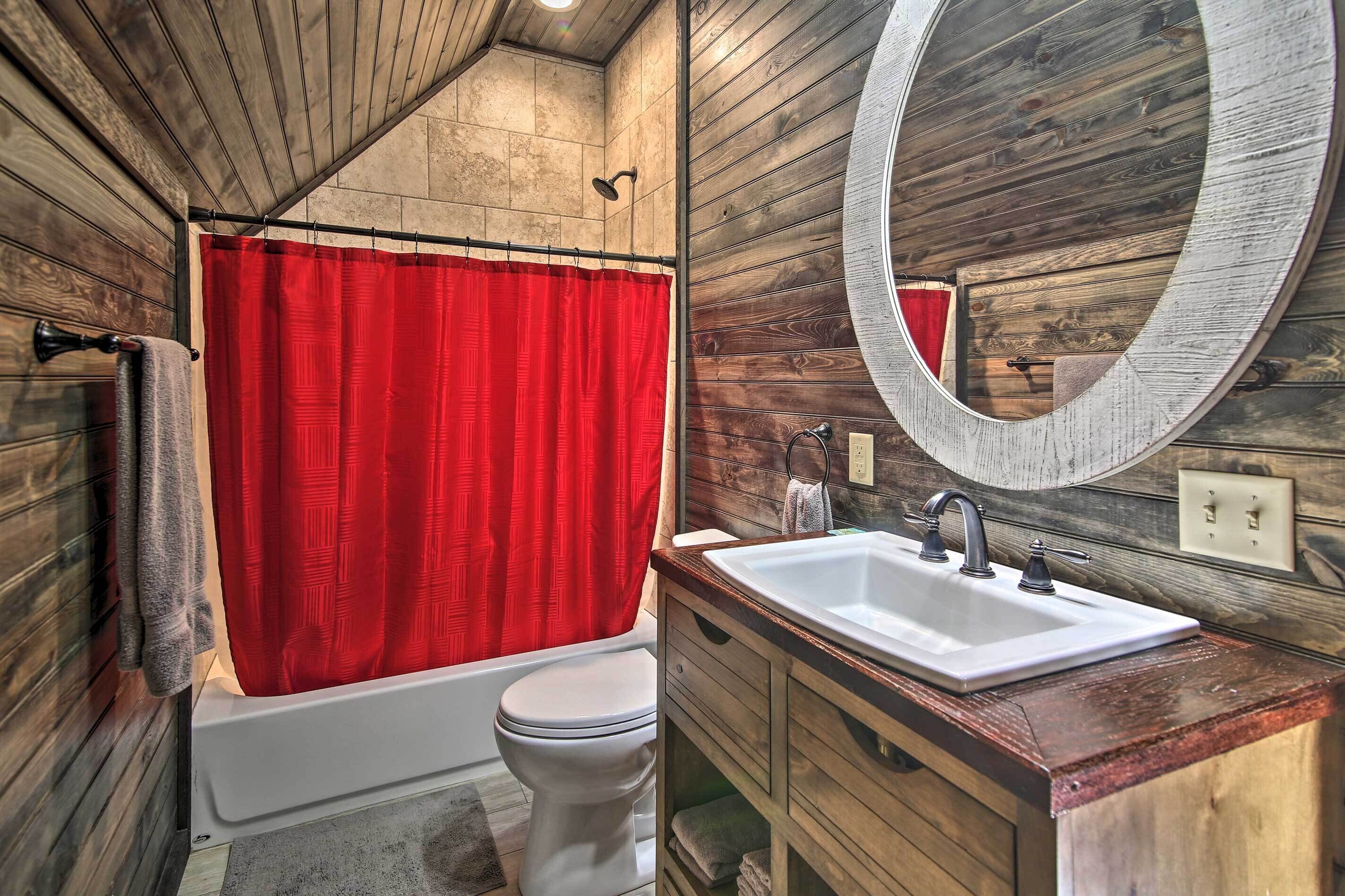 A bathroom with a wooden interior, a red shower curtain, a white bathtub, a white toilet, and a large mirror above the sink.