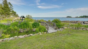 A wooden bridge over a rocky shoreline, a gazebo, and a grassy area with trees and shrubs. - Waterfront Prospect Harbor Cottage: Yard, Fire Pit (Prospect Harbor)