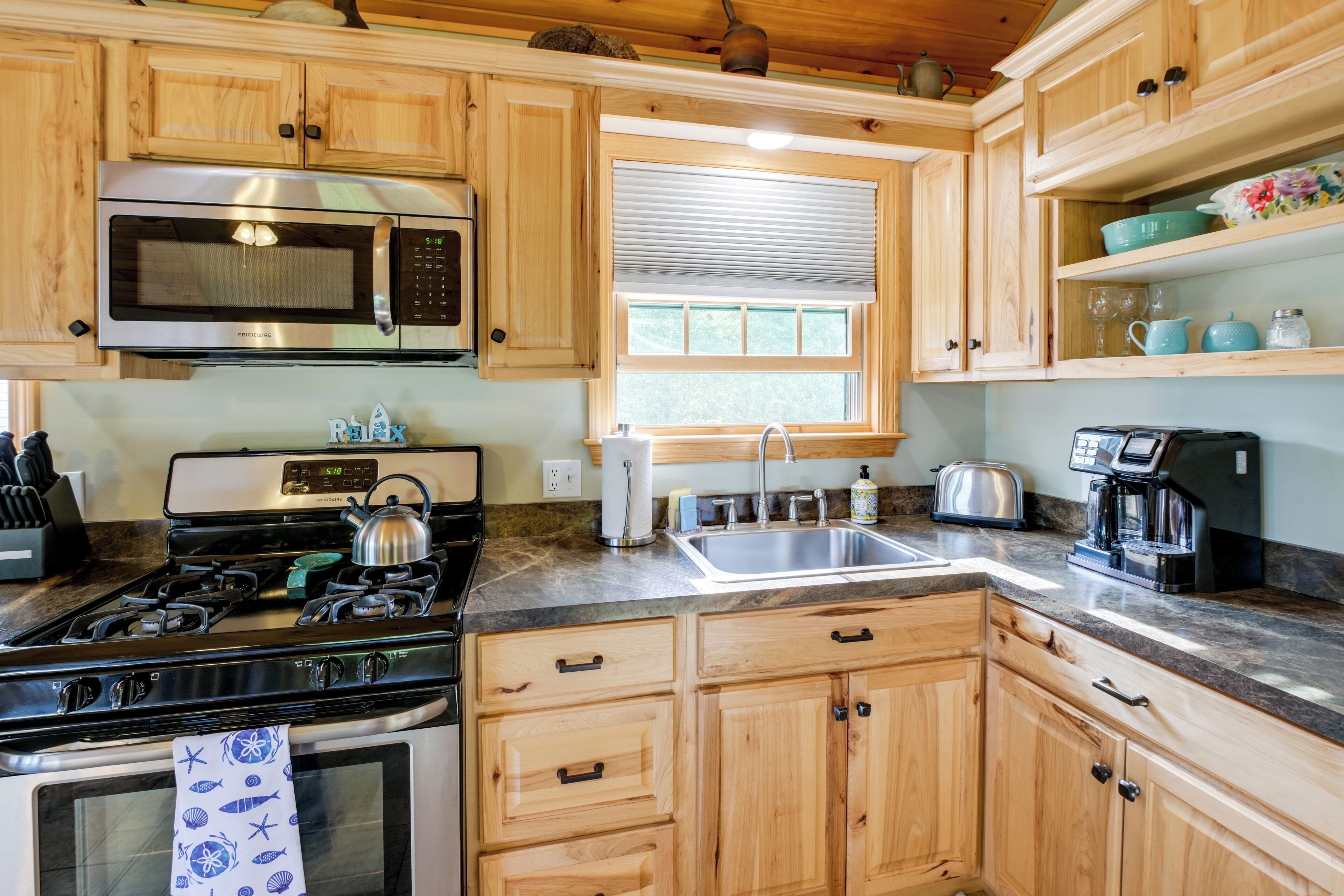 Cocina con gabinetes de madera, un microondas de acero inoxidable, estufa a gas, pileta y ventana con persianas.