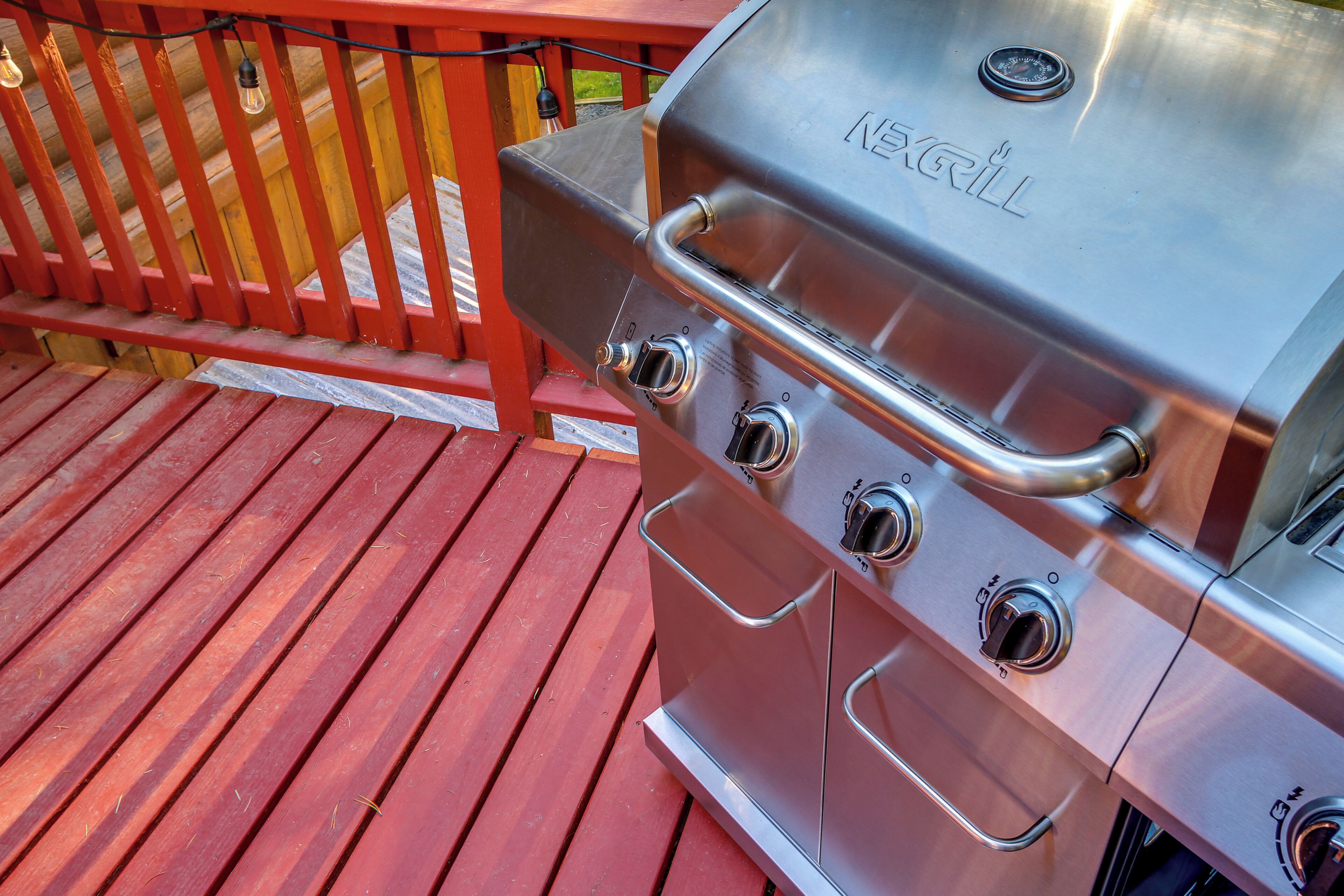A stainless steel outdoor grill with four burners and a control panel, placed on a wooden deck with a red railing.
