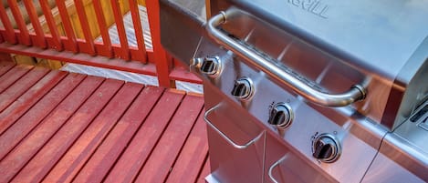 A stainless steel outdoor grill with four burners and a control panel, placed on a wooden deck with a red railing.