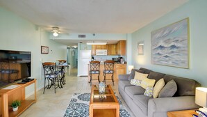A living room with a sofa, coffee table, and TV. There is a kitchen in the background with a microwave and cabinets.