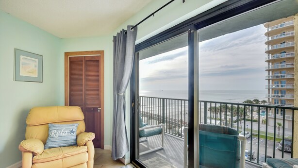 A balcony with a view of the ocean and a building, a yellow chair with a pillow, and a door with a curtain.
