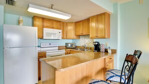 A kitchen with wooden cabinets, a white refrigerator, a microwave, a toaster, and a coffee maker. There are two chairs with patterned cushions.