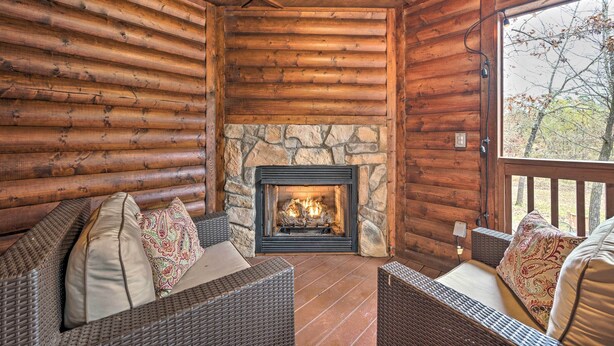 A log cabin interior with a stone fireplace, wicker furniture, and a view of trees outside. - Hot Tub: 'little Bear's Pond' Broken Bow Cabin (Broken Bow)