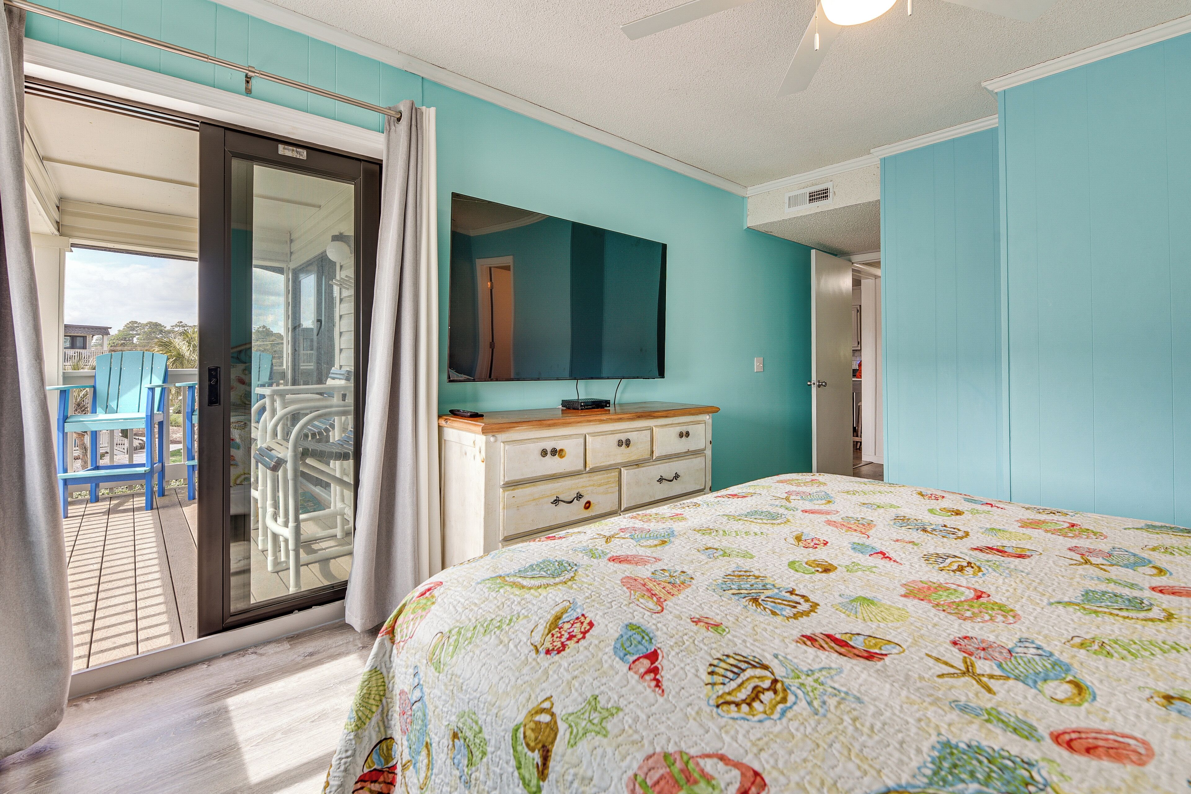 A bedroom with a large bed, a dresser, a mirror, and a sliding glass door leading to an outdoor area with blue chairs.