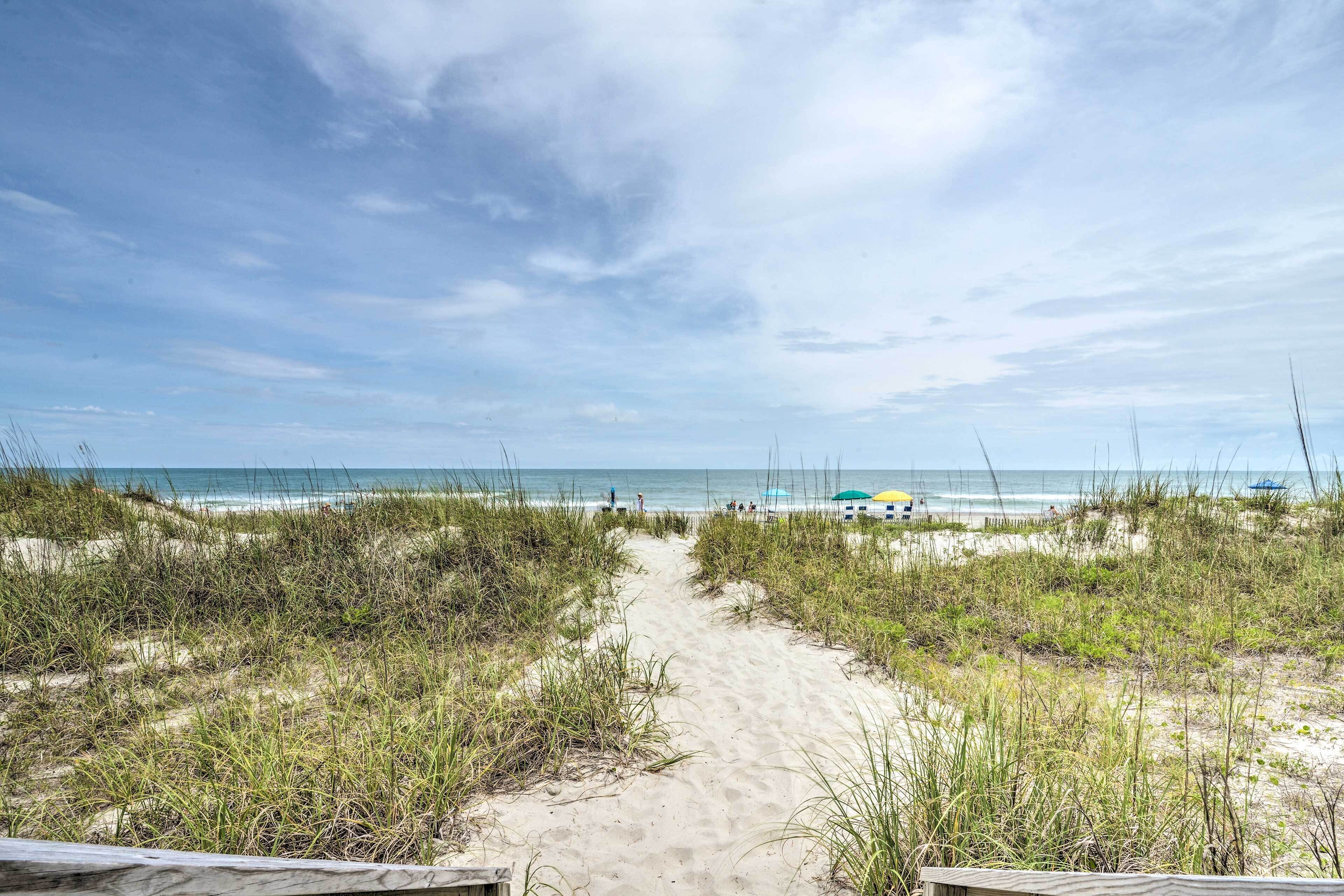 A sandy beach path leading to the ocean with dunes and beach umbrellas in the background.