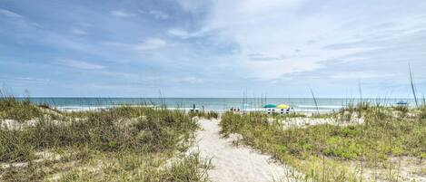 A sandy beach path leading to the ocean with dunes and beach umbrellas in the background.