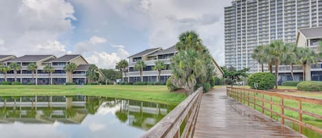 A wooden boardwalk over a calm pond, leading to a row of modern multi-story buildings with balconies.