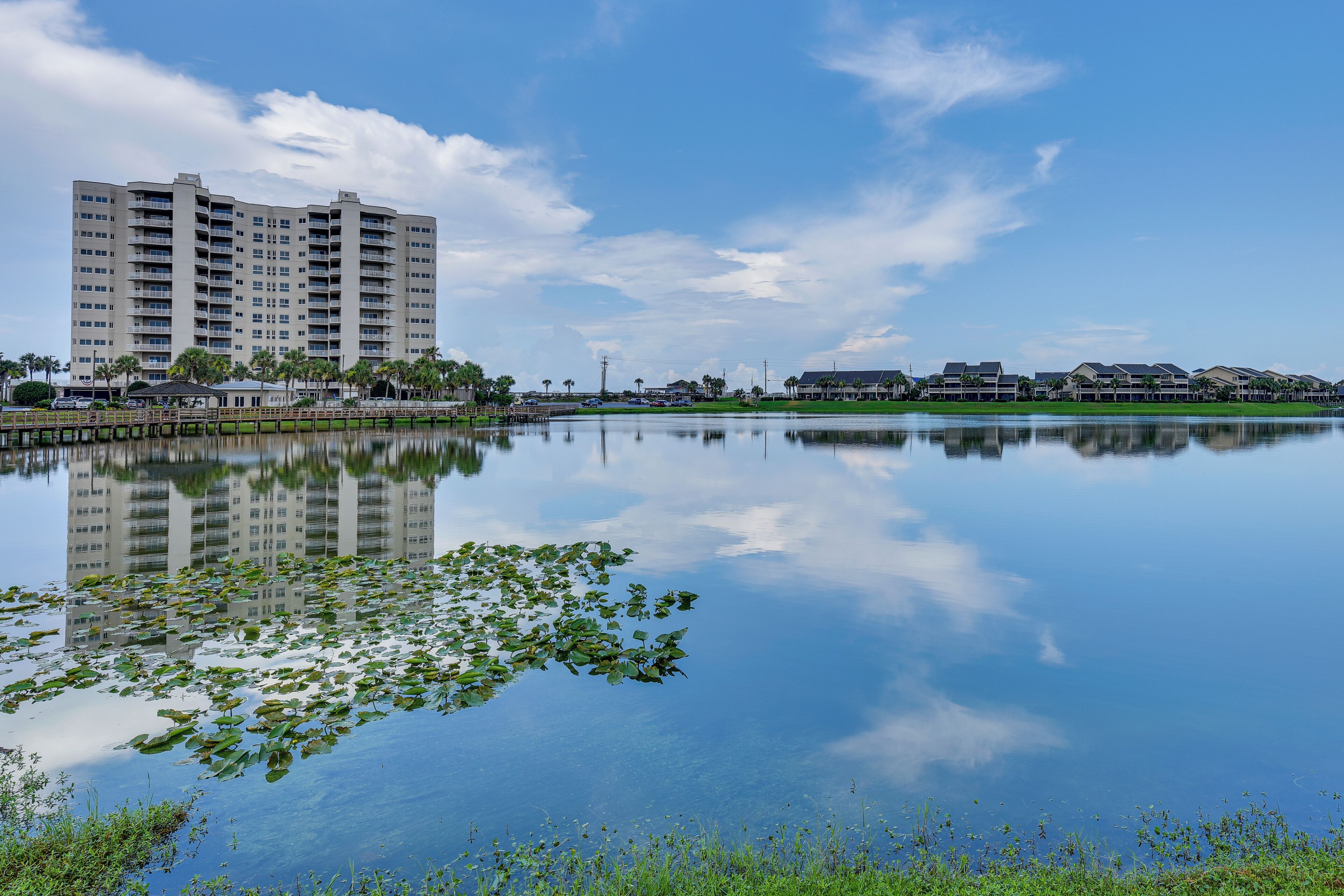 A large building with multiple floors and balconies, a calm body of water with lily pads, and a clear blue sky.